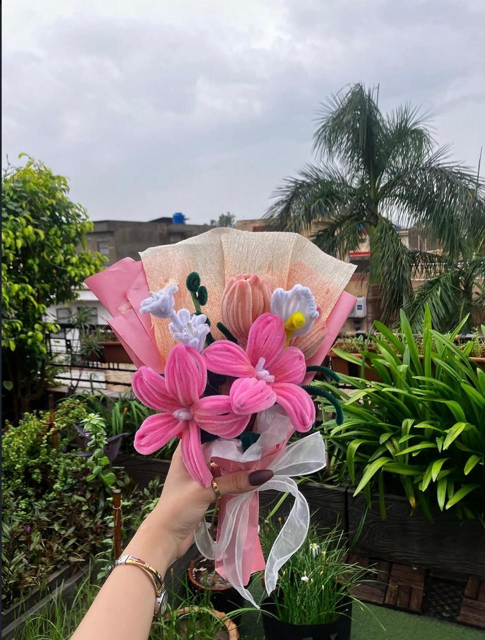 Bouquet of pink flowers held in front of a garden with greenery and buildings in the background