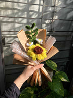 Hand holding a small bouquet of flowers with brown paper against a window with shadows.