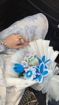Bouquet of flowers with blue and white decorations held by a person wearing a ring.