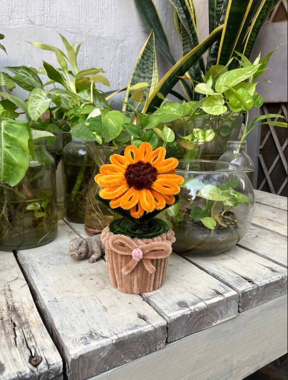 Decorative sunflower in a small pot on a wooden surface with other plants in the background.