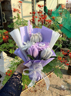 Bouquet of decorative flowers wrapped in purple paper with a white ribbon, held by a person in a garden setting.