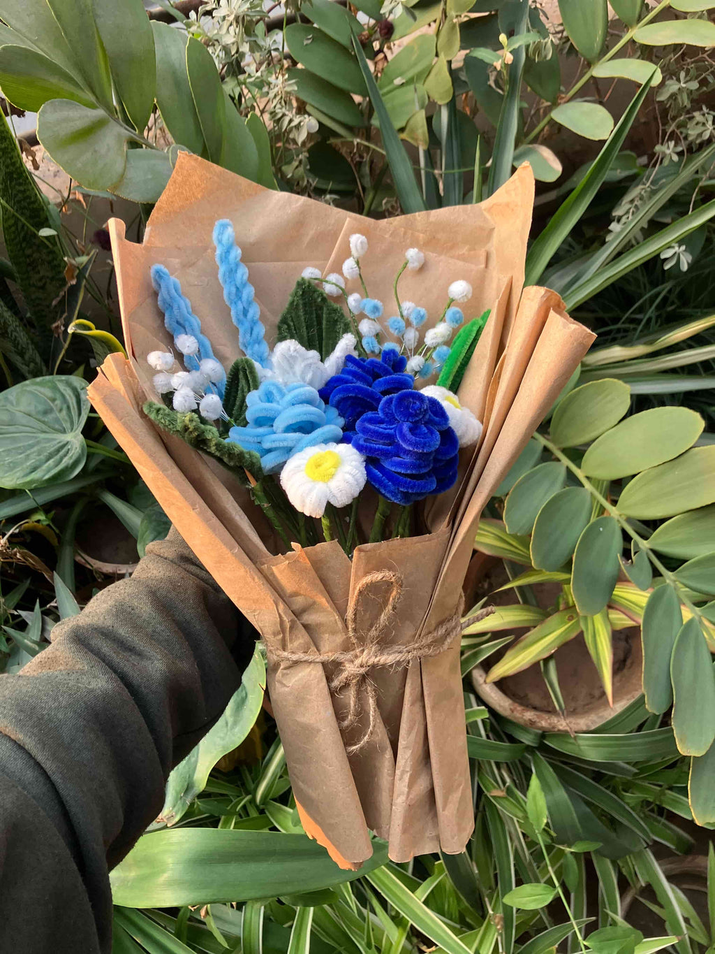 Bouquet of blue and white flowers wrapped in brown paper held by a hand against a green leafy background