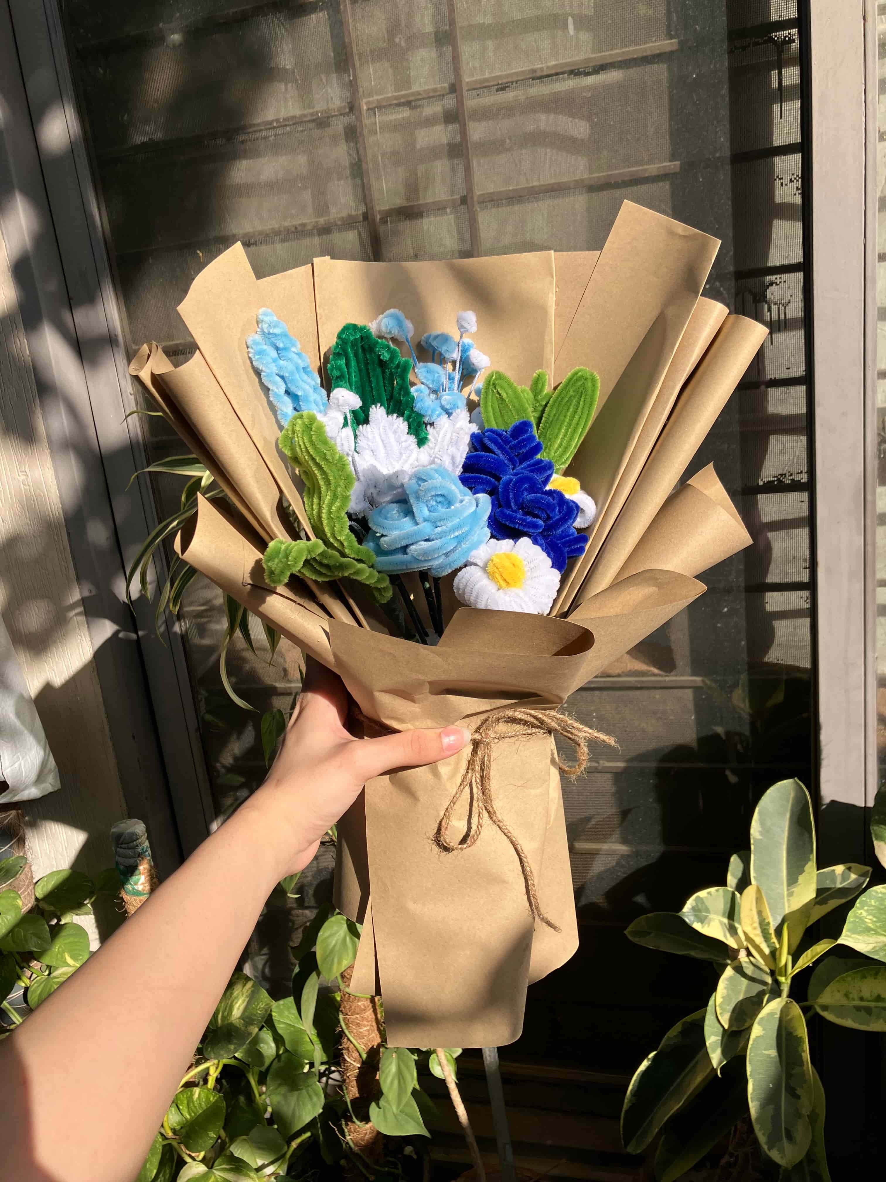 Bouquet of artificial flowers wrapped in brown paper on a glass table with plants in the background.