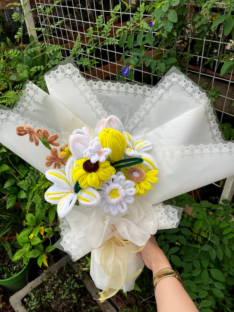 Knitted flower bouquet with white, yellow, and brown colors on a white lace-covered box against a garden background.