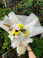 Knitted flower bouquet with white, yellow, and brown colors on a white lace-covered box against a garden background.