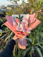 Bouquet of pink flowers wrapped in pink paper with a white ribbon, held against a natural background.
