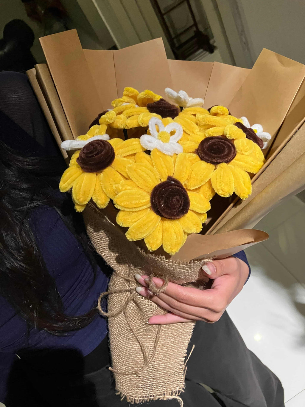 Hand holding a bouquet of yellow flowers with brown centers, wrapped in burlap, against a neutral background.
