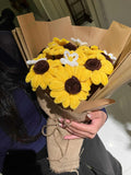 Hand holding a bouquet of yellow flowers with brown centers, wrapped in burlap, against a neutral background.