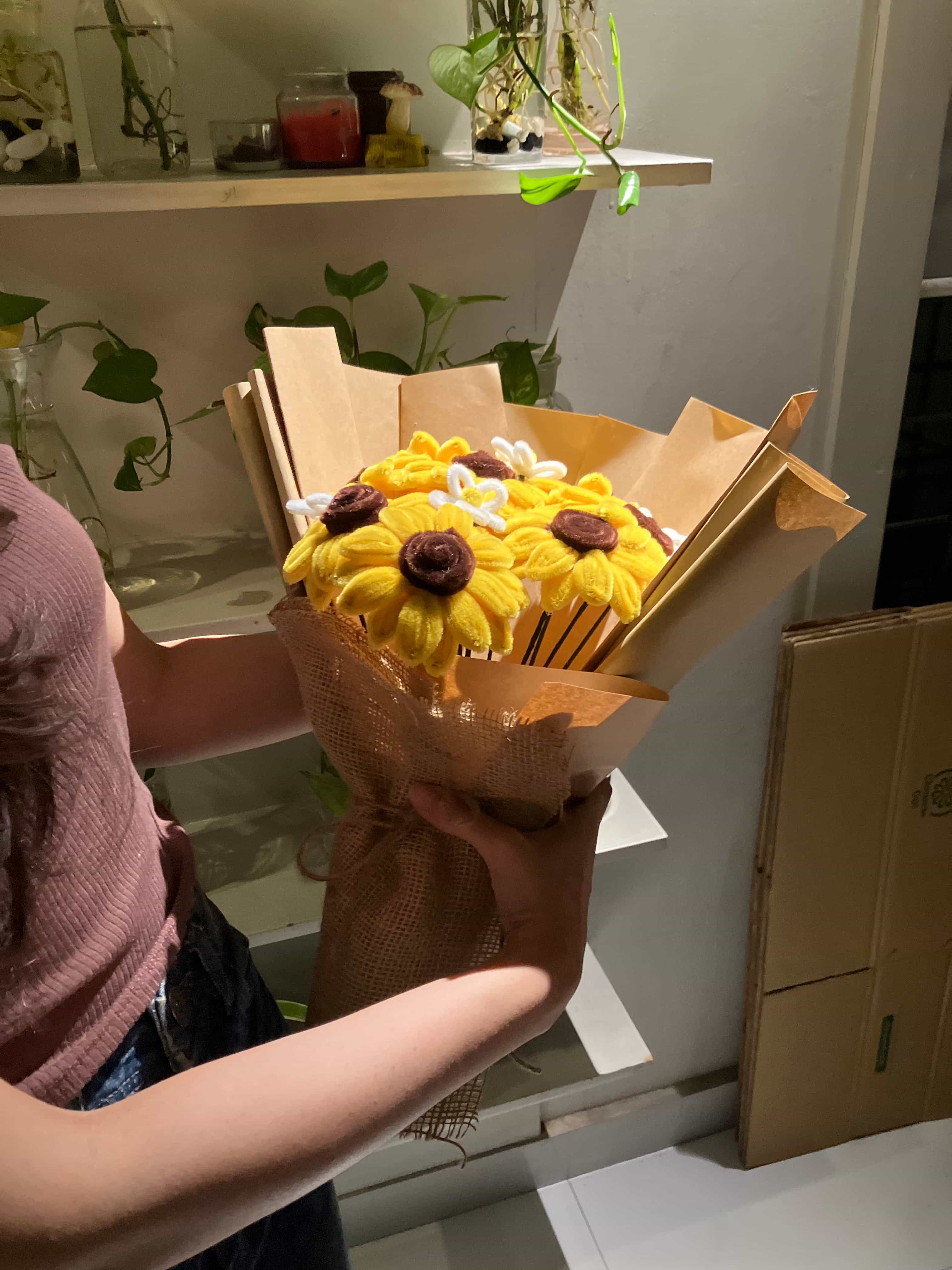 Person holding a decorative sunflower item in a room with plants and furniture.