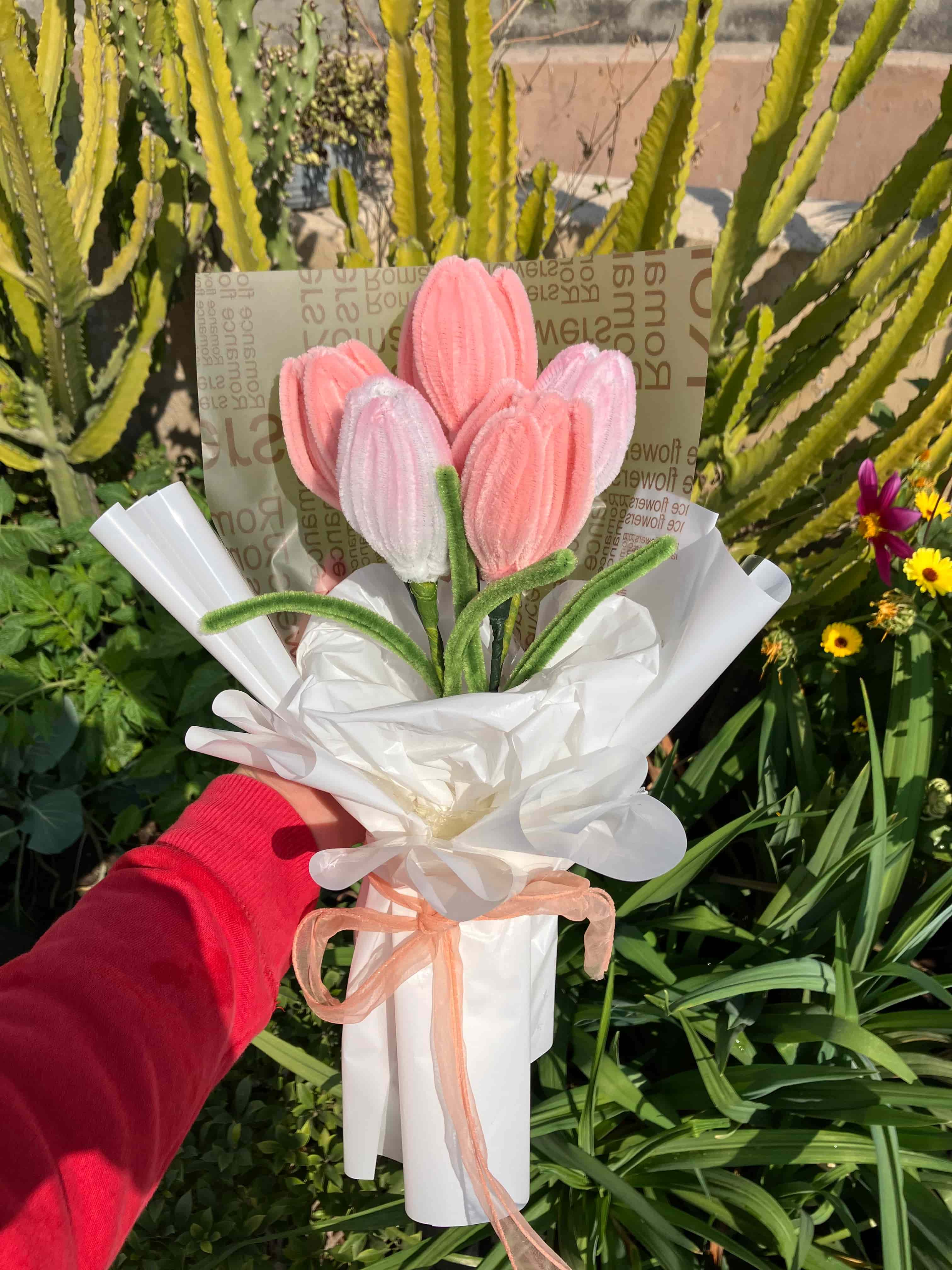 Bouquet of pink and white flowers held by a person wearing red gloves, with green plants and a newspaper in the background.