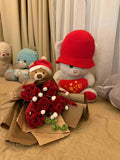Bouquet of red flowers with a teddy bear wearing a red hat on a beige surface.