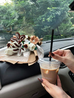 Person holding a coffee cup with a straw, sitting inside a vehicle with a blurred green outdoor background.