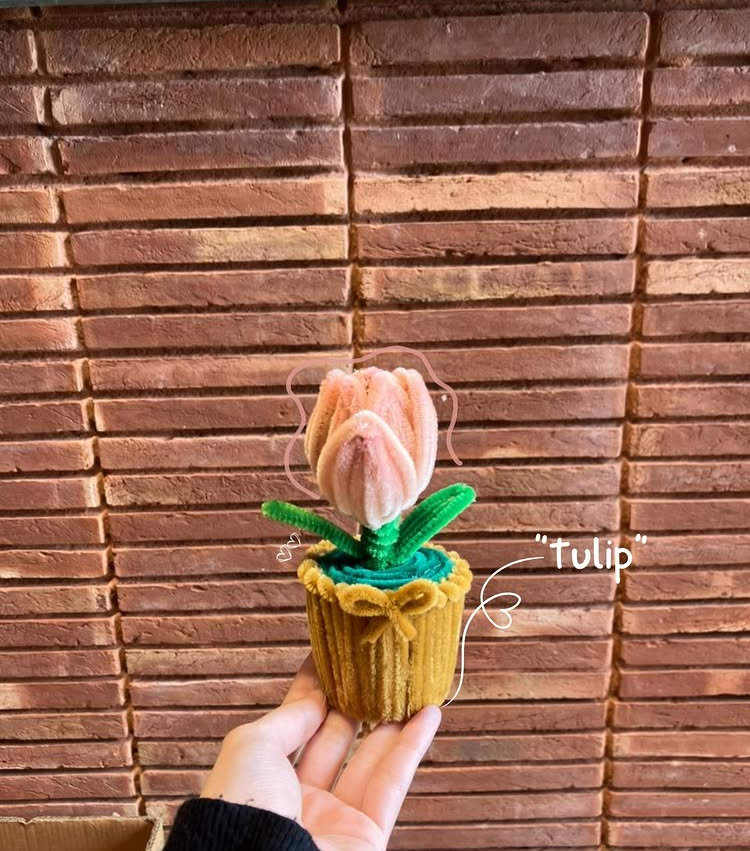 Hand holding a small knitted tulip plant against a brick wall.