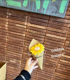 Hand holding a small yellow flower arrangement labeled 'Gerbera Bookev' against a wooden background.