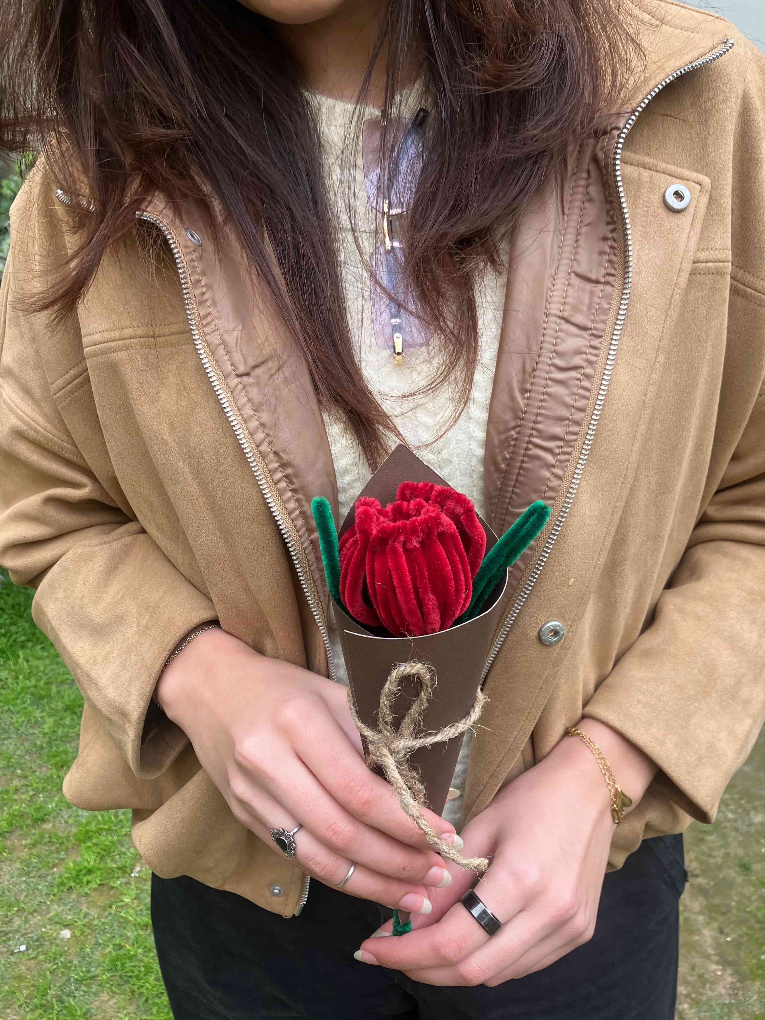 Person wearing a beige jacket with a red flower held in front of it, outdoors.