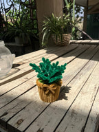 Artificial green plant in a brown pot on a wooden deck with plants in the background.