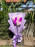 Bouquet of pink and purple flowers wrapped in purple paper with a bow, set against a garden background.