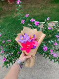 A hand holding a small wrapped bouquet of artificial flowers, including purple and pink blossoms, in front of a real garden with green foliage and pink flowers.