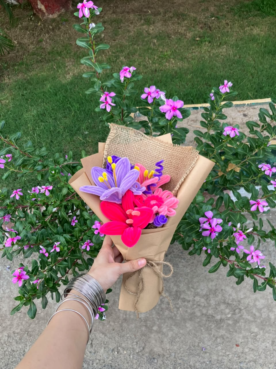 A hand holding a small wrapped bouquet of artificial flowers, including purple and pink blossoms, in front of a real garden with green foliage and pink flowers.