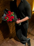 A person in a black shirt and black pants holding a large bouquet of red roses wrapped in black paper, standing on a wooden staircase in a dimly lit indoor setting.