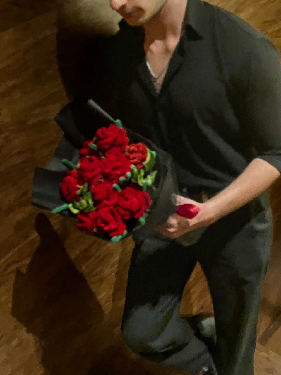 A person in a black shirt and pants holding a bouquet of red roses wrapped in black paper, standing on a wooden floor.