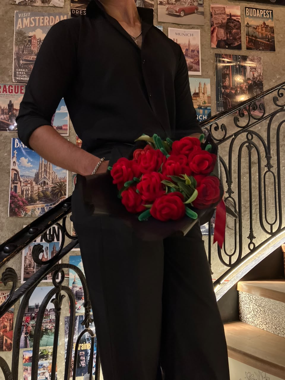 A person wearing a black button-up shirt and black pants holds a bouquet of red roses with green leaves in front of a staircase with a black metal railing and a wall covered in travel posters.