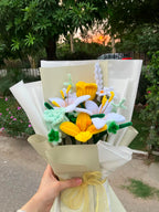 A handmade crochet flower bouquet held by a hand outdoors. The bouquet features crocheted white lilies, yellow daffodils, and a light green crocheted sprig, wrapped in cream-colored paper with a yellow ribbon.