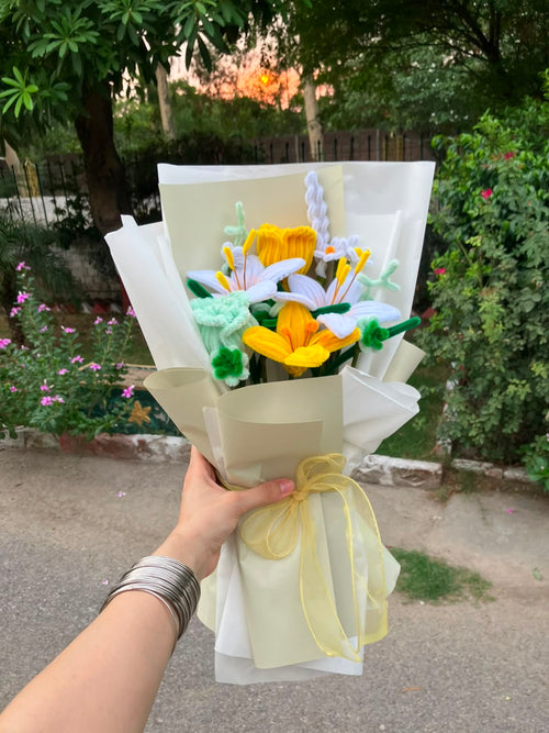 A hand holding a handmade crocheted flower bouquet with yellow tulips, white lilies, and green leaves, wrapped in cream paper with a yellow ribbon, set against an outdoor garden background.