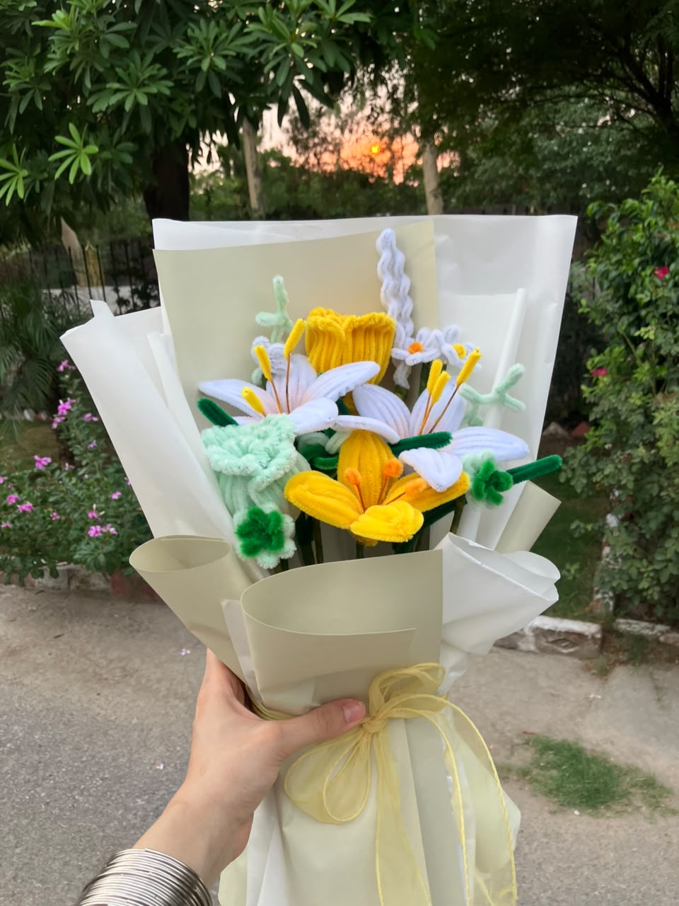 A handmade crochet flower bouquet wrapped in cream paper and tied with a yellow ribbon, held by a hand in an outdoor garden setting.