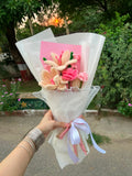 A handmade crocheted flower bouquet held by a hand, wrapped in white tissue paper with a pink card and a light purple ribbon, set against an outdoor garden background.
