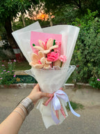 A handmade crocheted flower bouquet held by a hand, wrapped in white tissue paper with a pink card and a light purple ribbon, set against an outdoor garden background.