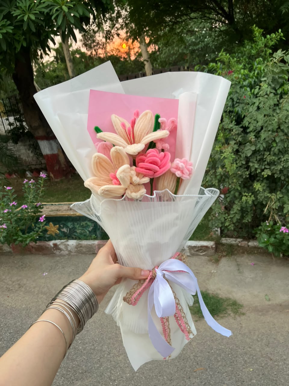 A handmade crocheted flower bouquet held by a hand, wrapped in white tissue paper with a pink card and a light purple ribbon, set against an outdoor garden background.