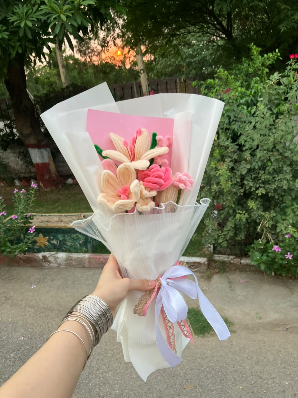 A hand holding a small decorative bouquet wrapped in white tissue paper, featuring pink and beige flower-like elements with a pink card insert and a white ribbon bow, set against an outdoor garden background.