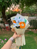 A handmade felt flower bouquet held by a hand in an outdoor garden setting. The bouquet features white daisy-like flowers with green stems, a bright orange daisy with a white center, and a small orange flower, all wrapped in beige paper with twine.