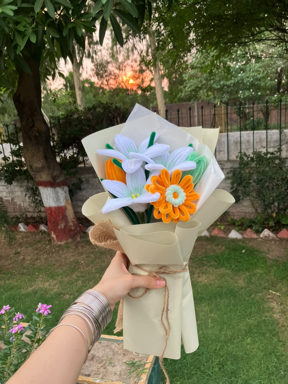 A handmade felt flower bouquet held by a hand in an outdoor garden setting. The bouquet features white daisy-like flowers with green stems, a bright orange daisy with a white center, and a small orange flower, all wrapped in beige paper with twine.